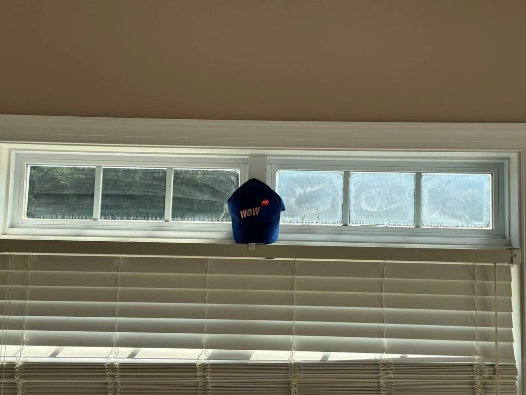Frosted basement window showing condensation issues with WOW FIX company hat, demonstrating common window repair needs in North Carolina