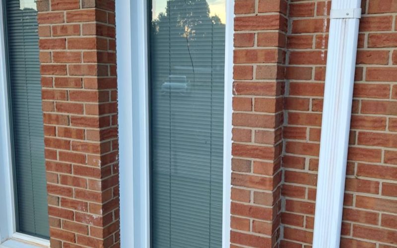 Cracked full-length window with white trim in brick home exterior, showing residential window damage needing professional repair