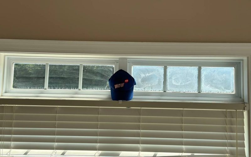 Frosted basement window showing condensation issues with WOW FIX company hat, demonstrating common window repair needs in North Carolina