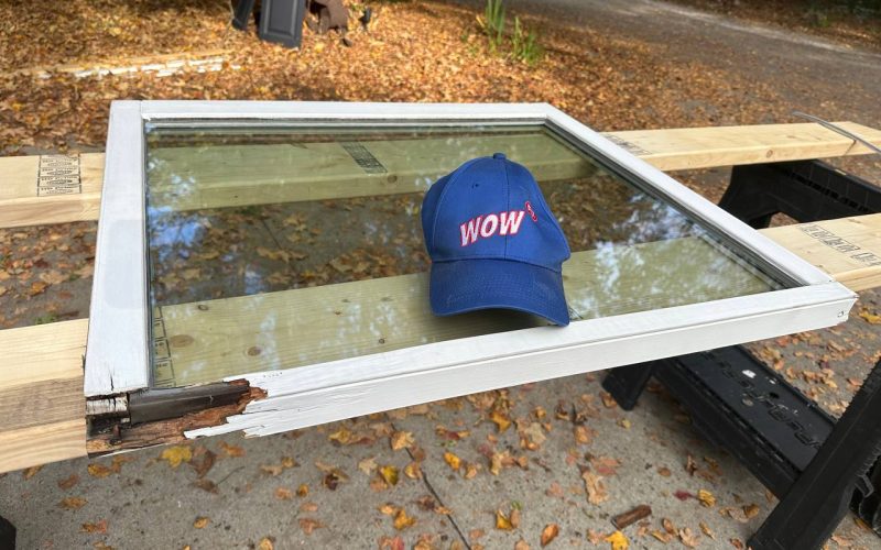 Damaged white window frame with WOW company hat showing wood rot damage, demonstrating before condition for window repair service