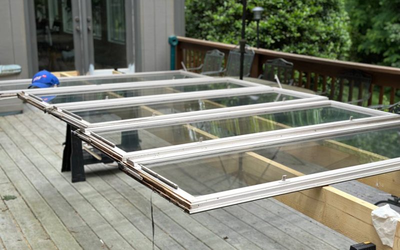 Window sash repair and restoration in progress showing multiple white-framed glass panels laid out on sawhorses on wooden deck