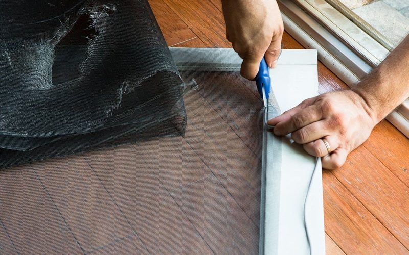 Professional window screen repair technician cutting new mesh material for residential window screen replacement
